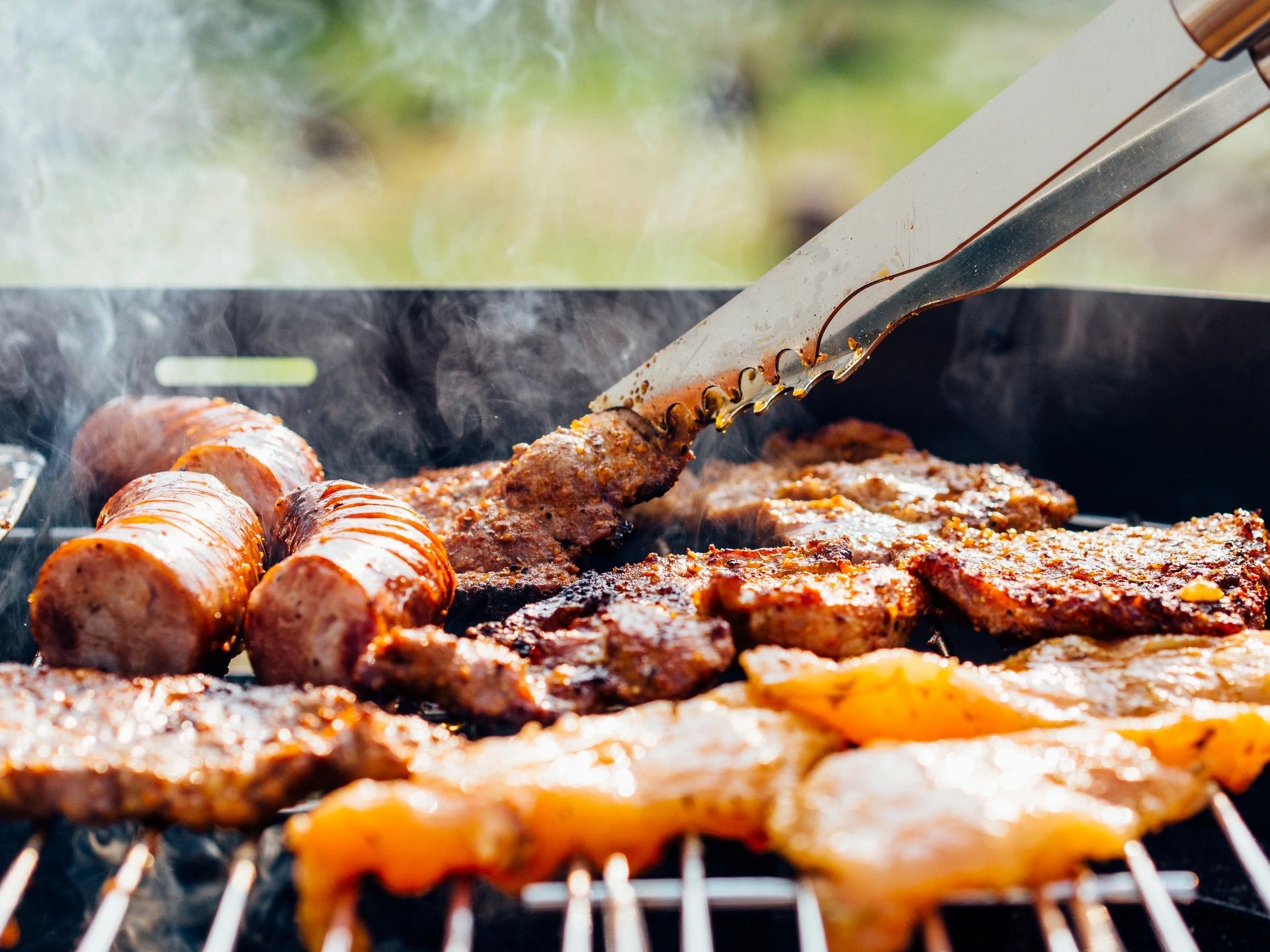 Grilled meat sizzling on an outdoor barbecue grill with tongs.